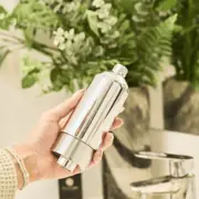 Hånd der holder et cylinderformet brusefilter i blank krom foran grønne planter
